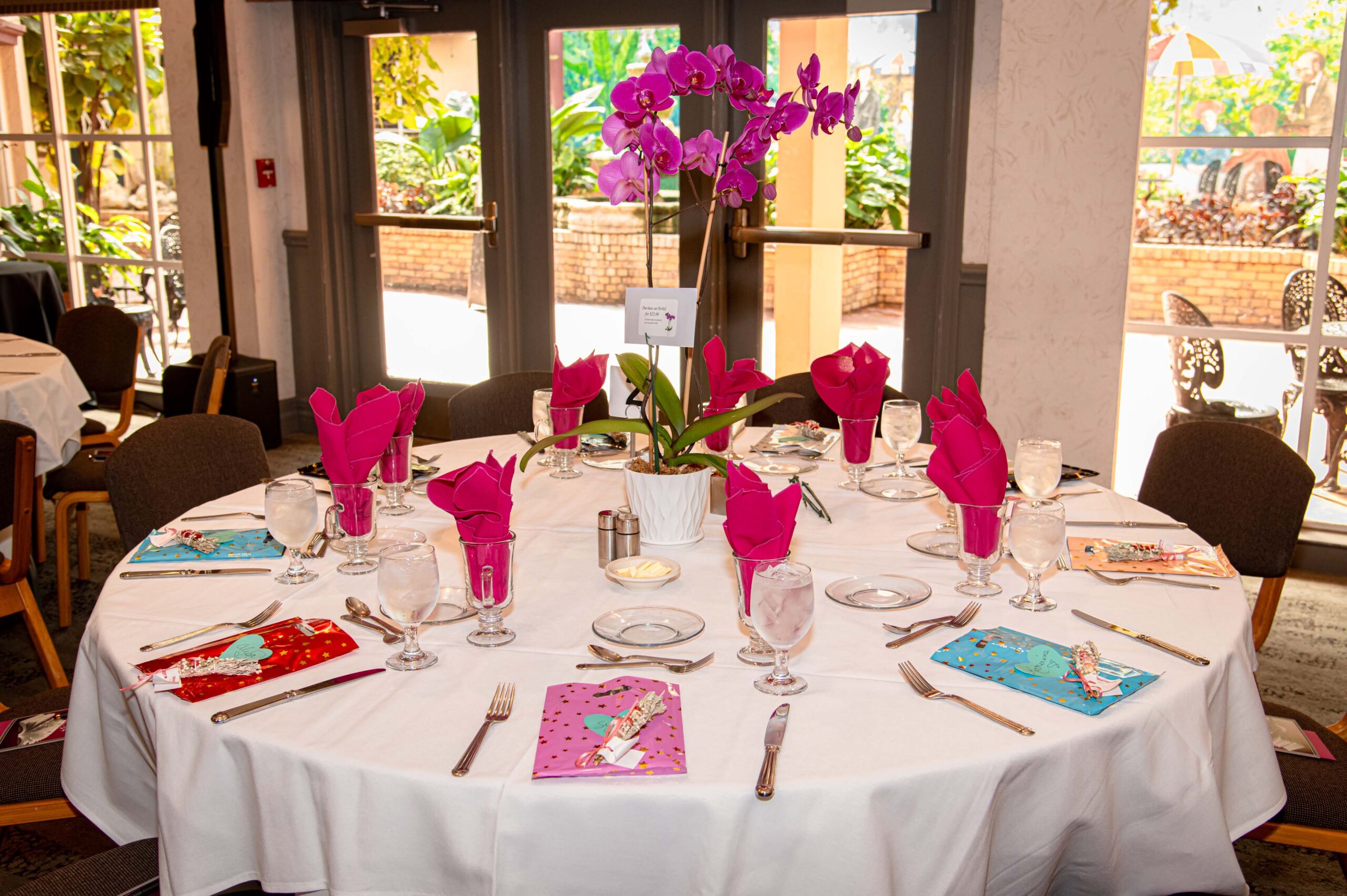 a table next to a window with colorful bags at each place setting, glasses with red napkins and a tall pink orchid